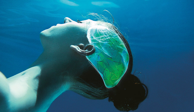 A woman floats under water with her eyes closed. Only her neck and head are depicted. On the side of her head is a graphic in the shape of a brain. The back part of the graphic is green, while the front part is turquoise.